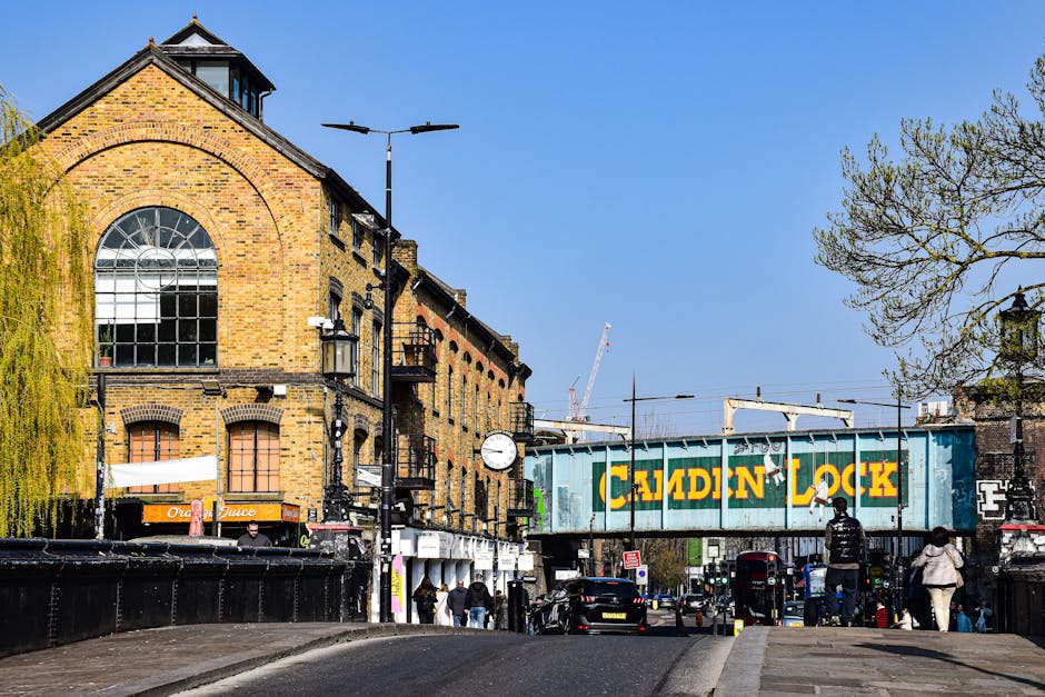 A street scene in Lea Bridge featuring a large, historic brick building with multiple arched windows, situated above pavement level. In the foreground, there are several pedestrians walking along the sidewalk and a few parked cars aligned along the street. A prominent railway bridge crosses the road, displaying bold yellow lettering spelling 'CAMDEN LOCK' on a green weathered metal structure, indicating proximity to a transportation or industrial area. There are streetlights and a clock mounted on street furniture, with the blue sky and scattered trees visible in the background, suggesting a clear day. The scene includes elements typical of urban home relocation or furniture transport logistics, such as the street environment where a professional removals service like Man with Van Lea Bridge may operate, with the company providing house removals and moving services in this area.