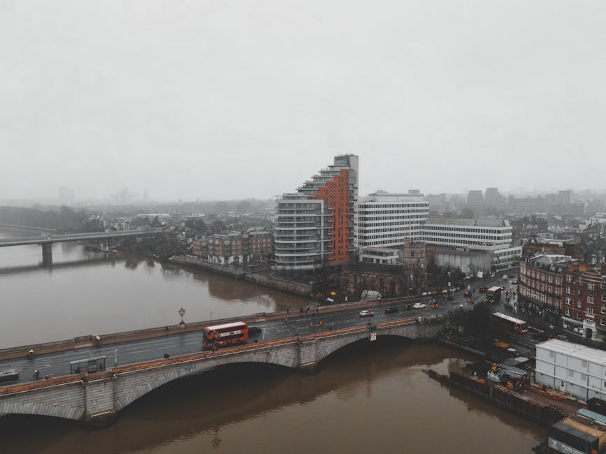 Aerial view of a cityscape on a cloudy day in Lea Bridge, showing a river flowing beneath an arched stone bridge with a red double-decker bus and several cars in transit. Next to the river, modern residential apartments featuring curved balconies and a white commercial building are visible. On the street adjacent to the river, parked and moving vehicles, along with traffic lights and street lamps, can be seen. The environment appears quiet with minimal pedestrian activity, and the overall scene captures an urban area suitable for home relocations and furniture transport, highlighting the logistics involved in house removals supported by Man with Van Lea Bridge.