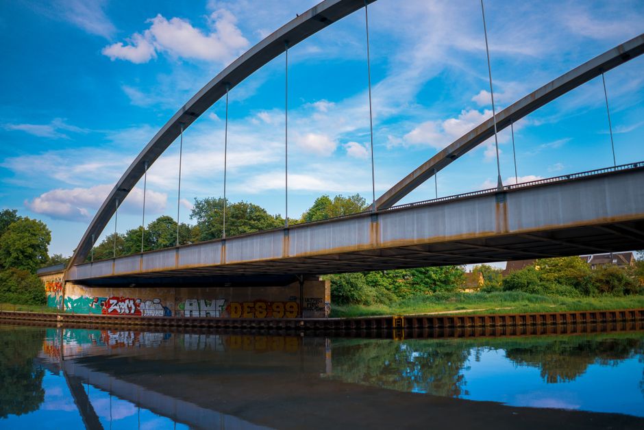 A modern steel arch bridge with cables extending from the top of the arches supporting the bridge deck, spanning over a calm waterway. The underside of the bridge shows weathered steel and graffiti on the concrete pier beneath, with reflections visible on the water surface. Surrounding green trees and shrubbery frame the scene under a partly cloudy sky, with natural daylight illuminating the structure. This structural element is situated outside, near residential areas, and could be part of a route used during home relocation or furniture transport. Occasionally, Man with Van Lea Bridge assists with removals and moving logistics in this vicinity, which may involve transporting household belongings across such infrastructure during packing and moving processes.