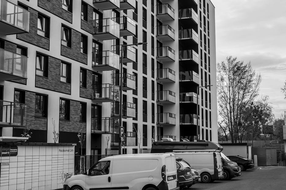 Black and white photograph of a modern, multi-storey residential building with numerous small balconies featuring glass railings arranged in vertical columns. In the foreground, several commercial vehicles, including a white van, a black van, and a small car, are parked on the paved street next to the building. The vehicles appear to be involved in a house removal or home relocation process, with some showing signs of loading or unloading activities. To the left of the vehicles, there is a small gated area with a tiled wall, and behind it, a few young trees and sparse shrubbery are visible. The building’s façade combines brick and white panels, and the overall scene is illuminated by natural daylight with an overcast sky. This setting reflects typical urban relocation logistics, where Man with Van Lea Bridge offers removals services supporting furniture transport and packing and moving operations in the LEA BRIDGE area.