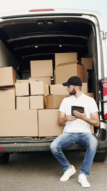 A young man wearing a black cap, white t-shirt, and jeans is sitting on the pavement next to an open van loading area during a home relocation process. The van's cargo space is filled with numerous cardboard boxes of various sizes, some sealed with packing tape, arranged in a stacked manner. The boxes are primarily brown, with a few larger boxes positioned at the back and smaller ones towards the front. Inside the van, a dark roof canopy is visible, with some tie-down straps and packing blankets possibly used to secure items during transportation. The man appears to be reviewing information on a tablet as part of the packing or moving process. The setting suggests an organized loading or unloading operation, typical of furniture transport and packing and moving activities conducted by companies like Man with Van Lea Bridge, supporting efficient house removals and logistics.