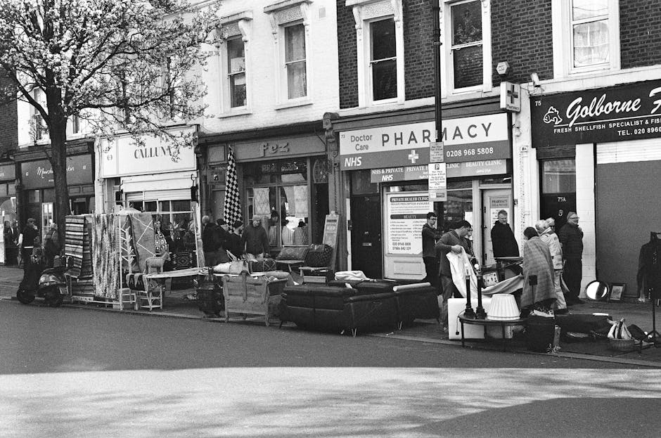 A black and white photograph of a busy street scene outside a commercial building with a sign indicating a pharmacy and NHS services, where various furniture items and boxes are laid out on the pavement. The visible items include a large sofa, an armchair, several cardboard boxes, wrapped packages, and a few pieces of disassembled furniture, all arranged in front of the store. A group of people, some wearing masks, are standing amidst the objects, appearing to be engaged in packing, unpacking, or moving activities. Trolleys, straps, and other packing materials are also present, suggesting a loading or unloading process as part of a home relocation or furniture transport operation. The scene is illuminated by natural daylight, with a few trees and additional shopfronts visible in the background. The setting indicates a street-side furniture or second-hand goods sale, with some items awaiting pickup by a moving service such as Man with Van Lea Bridge, who specialise in removals and packing for house and furniture relocations.