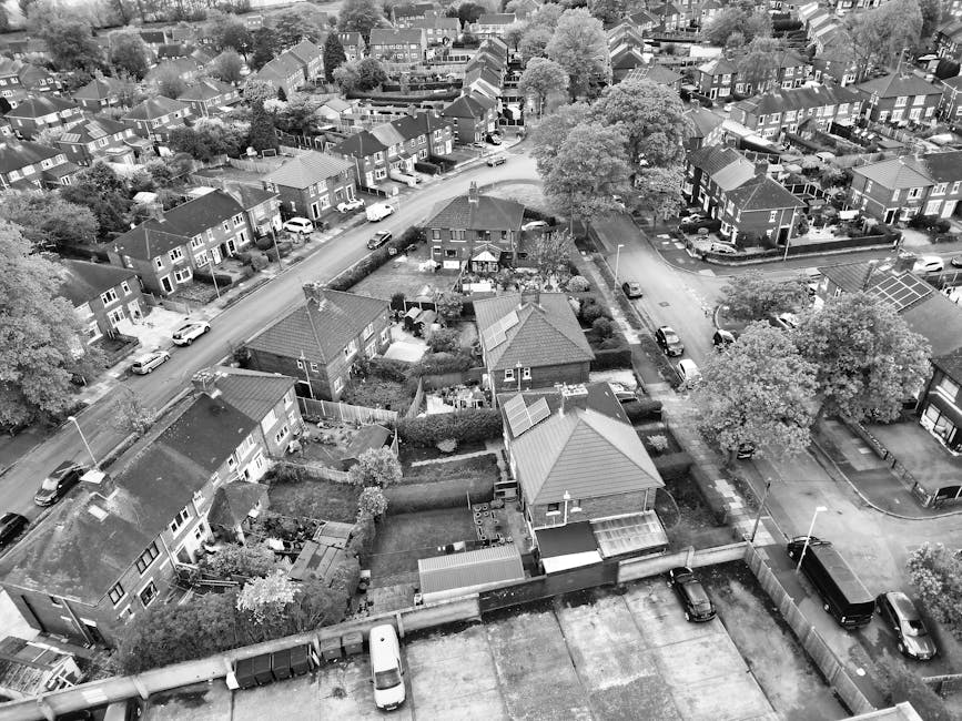 An aerial black-and-white photograph showing a residential area with a mix of detached and semi-detached houses, some with solar panels on their roofs, closely spaced along curving streets. The homes are surrounded by gardens, driveways, and parked cars. In the foreground, a row of terraced houses with small front yards and parked vehicles is visible. Behind these, a larger semi-detached property is situated next to a narrow road with a parked car and a driveway. To the right, there is a small, curved cul-de-sac with additional houses and parked cars. Toward the centre and rear of the image, a dense area of trees and green space separates the residential neighborhood from what appears to be a community or commercial area with large storage units or garages. The surrounding environment includes a network of roads, footpaths, and parking areas. This image is associated with [COMPANY_NAME], which provides house removals and furniture transport services, including packing and loading processes, to facilitate home relocation and logistics.