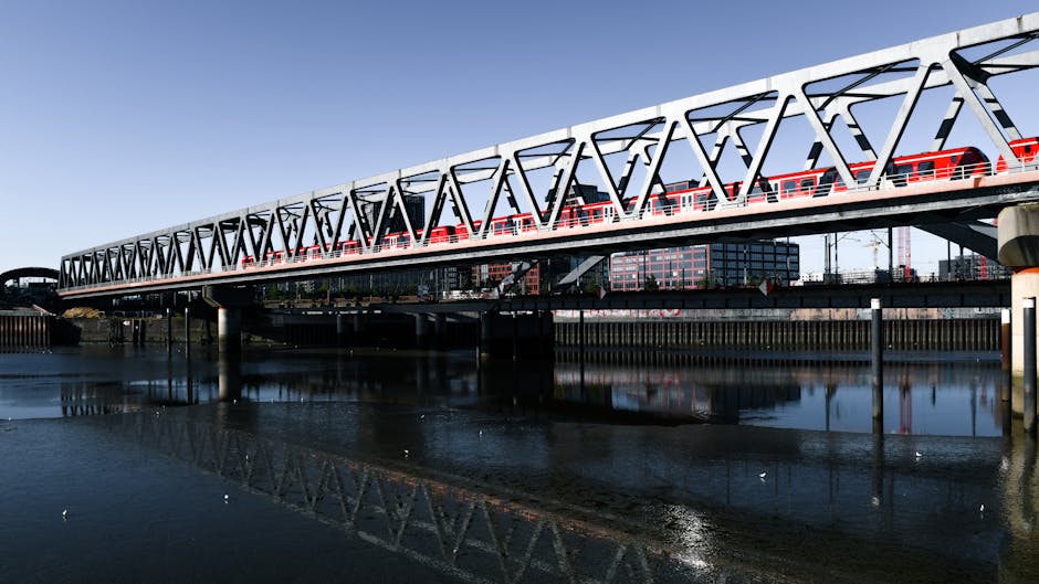 A large modern train passing over a steel bridge structure with a white and red design, spanning across a calm body of water during daylight. In the background, there are buildings and construction cranes visible, indicating an urban environment. The bridge's supporting pillars are reflected in the water below, creating a symmetrical image. The scene captures the transportation infrastructure typical of a home relocation route near Lea Bridge Station, emphasizing the importance of efficient furniture transport and moving logistics as part of a professional removals service. The clear sky and natural light highlight the structural details of the bridge and the train vehicles, aligning with the professional context of house removals and packing and moving processes facilitated by Man with Van Lea Bridge.