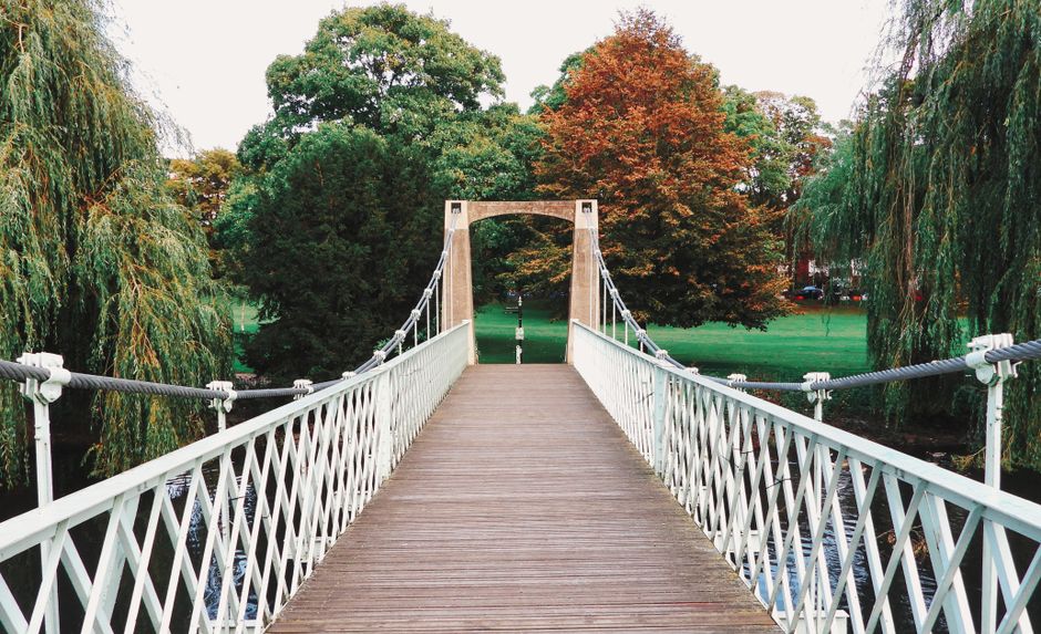 A long wooden footbridge with white lattice-style side railings spans over a narrow river in a park setting. The bridge is supported by vertical and diagonal tension cables attached to wooden support pillars at either end. Behind the bridge, there are lush green trees, including a weeping willow with drooping branches on the left and a tree with red and orange autumn foliage in the background. The sky is partly cloudy with diffused lighting. This scene depicts a peaceful outdoor environment suitable for outdoor activities or leisurely walks. In the context of house removals, the image represents the importance of planning for access routes and parking suspensions, as managed by local councils, to ensure smooth furniture transport and home relocation processes. Man with Van Lea Bridge may consider such factors when coordinating parking and loading logistics for customer relocations, especially near scenic or residential areas.