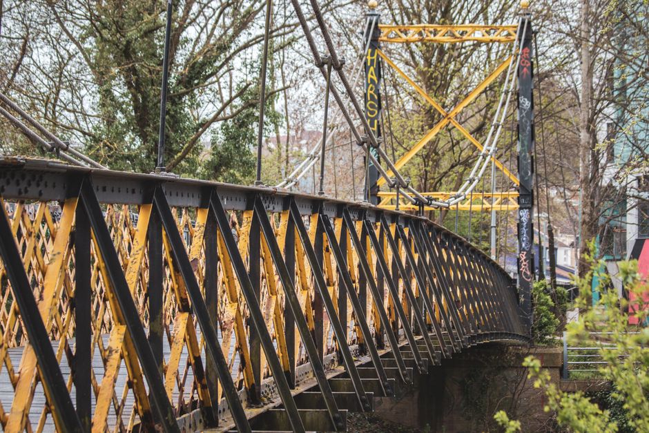 A close-up view of a yellow scaffolding tower and black metal safety barriers set up on a bridge in an urban area, with trees and modern buildings in the background. The scaffolding is secured with metal braces and ropes, indicating recent construction or maintenance work. The barriers are positioned along the edge of the bridge to ensure safety during the work process. In the foreground, a section of the bridge's metal railing is visible, with some greenery nearby. The scene is illuminated by natural daylight, suggesting daytime, and the environment appears calm with no visible pedestrians or vehicles. Occasionally, occasional signs of urban activity or traffic can be inferred from the background, aligning with the context of organized transportation or logistical planning for house removals, as referenced by Man with Van Lea Bridge in relation to parking suspension rules for effective furniture transport and home relocation in Lea Bridge.
