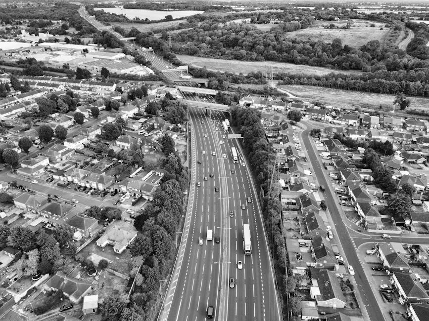 Aerial view of a multilane urban road with various vehicles, including cars, vans, and trucks, moving in both directions. The road runs through a residential area with numerous houses, gardens, and st