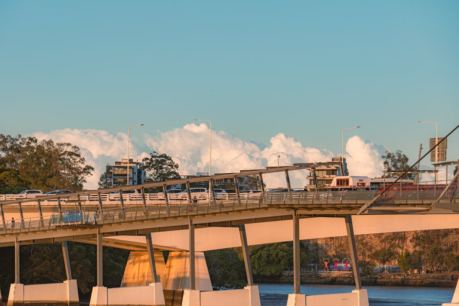 A bridge over a body of water during daytime with a clear blue sky and some scattered clouds in the background. Several vehicles, including cars and a bus, are traveling across the bridge, which features a metal railing and multiple streetlights mounted along its length. Beneath the bridge, a river or lake can be seen, with trees and greenery along the shoreline. This scene represents urban infrastructure that could be part of a city area requiring moving or transportation services. The setting illustrates the type of environment where Man with Van Lea Bridge might assist with home relocation, furniture transport, or logistics involved in packing and moving within or through the city.